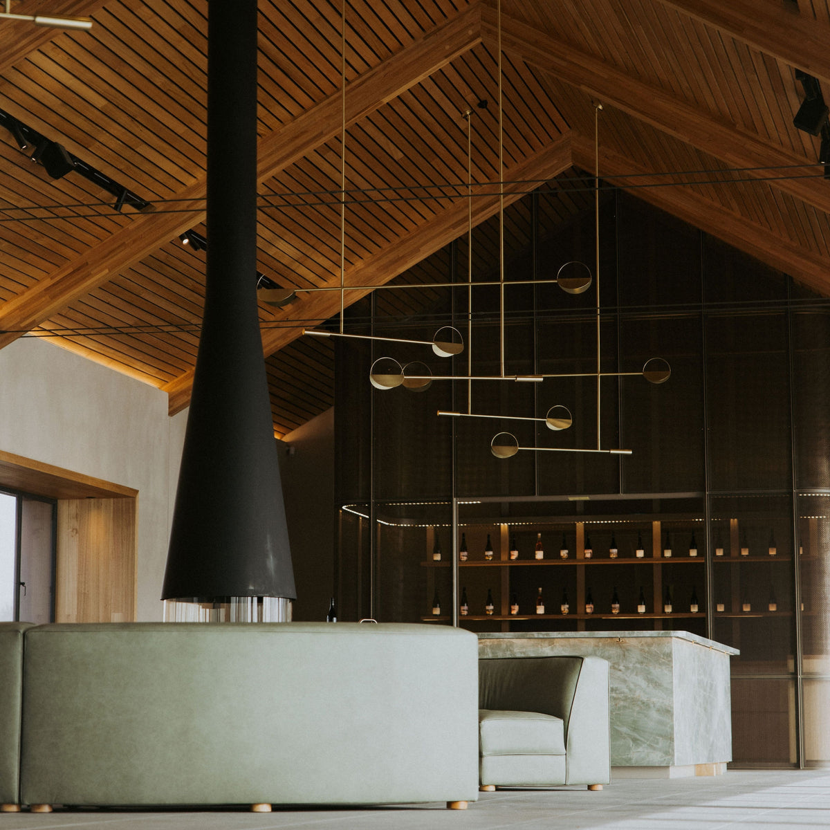 A sneak peek into our new Cellar Door & Restaurant with a large black conical fireplace or stove centered in the foreground, a pale green upholstered sofa, a marble-topped counter, and shelving in the background under a high wood-paneled roof.